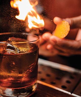 A glass of liquor sits on a bar as a bartender lights a flame.
