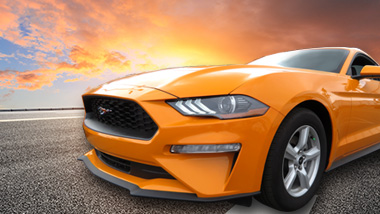 An orange Ford Mustang pictured on a paved road with a sunset behind it.